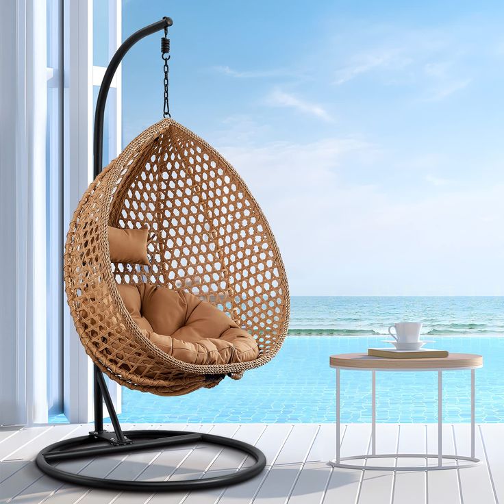 a brown hanging chair next to a table with a cup on it and the ocean in the background