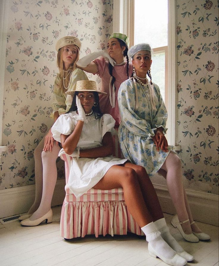 four women in dresses and hats are sitting on a bench near a window with floral wallpaper