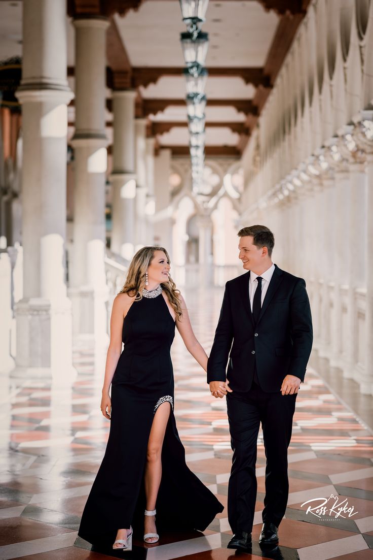 a man and woman in formal wear holding hands while walking through an ornate hallway with columns