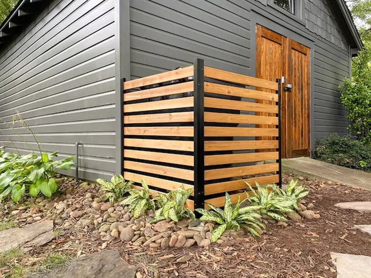 a wooden fence is in front of a gray building with green plants and rocks on the ground