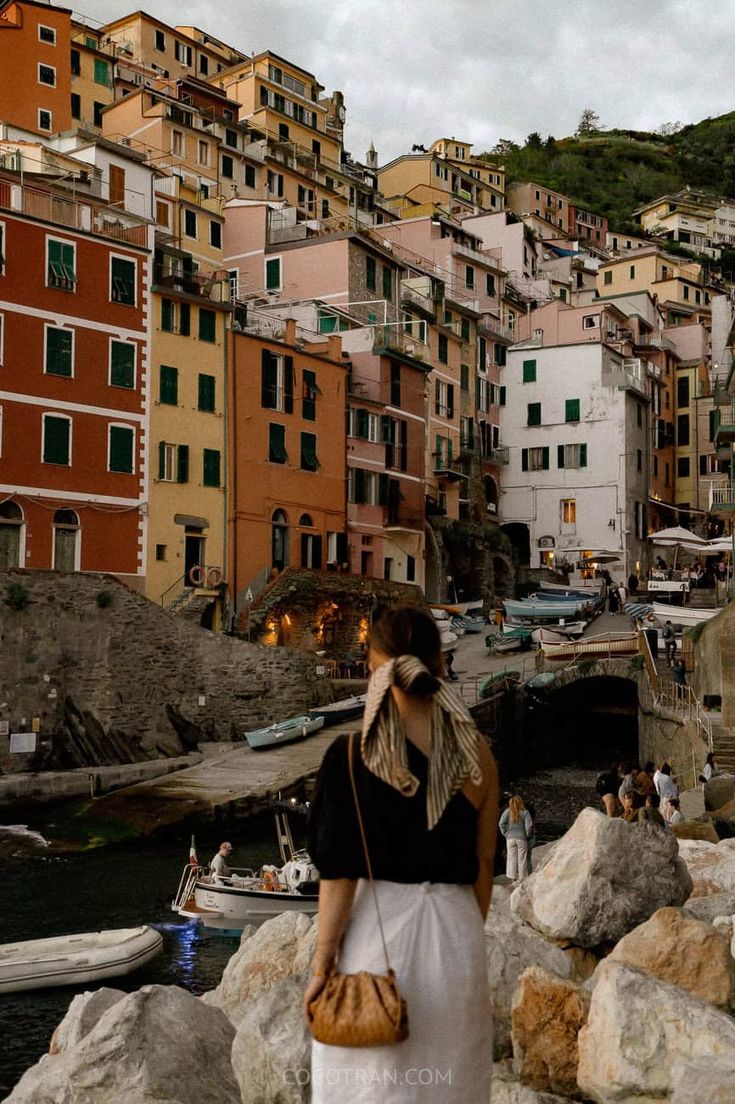 a woman is standing on the rocks looking at some boats in the water and buildings behind her