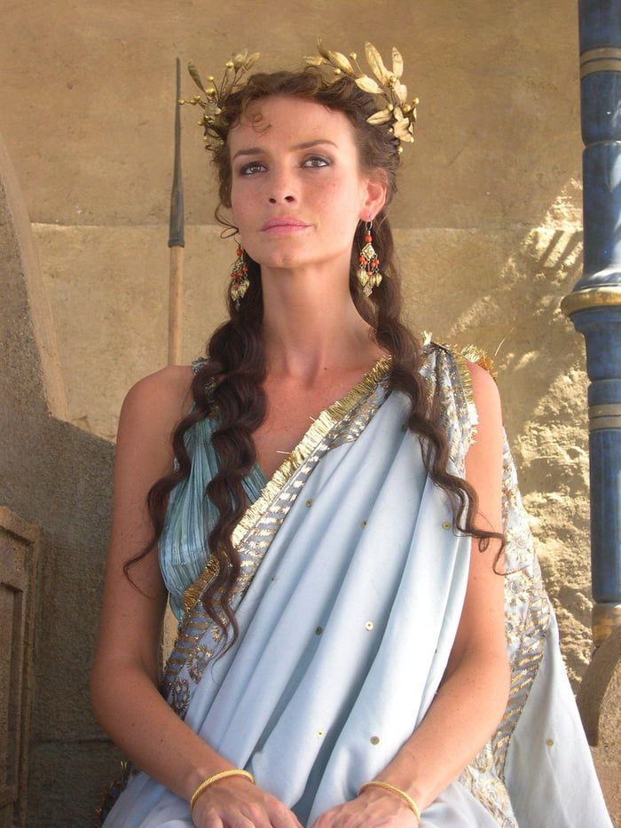 a woman dressed in an ancient greek costume with gold jewelry on her head, sitting against a stone wall