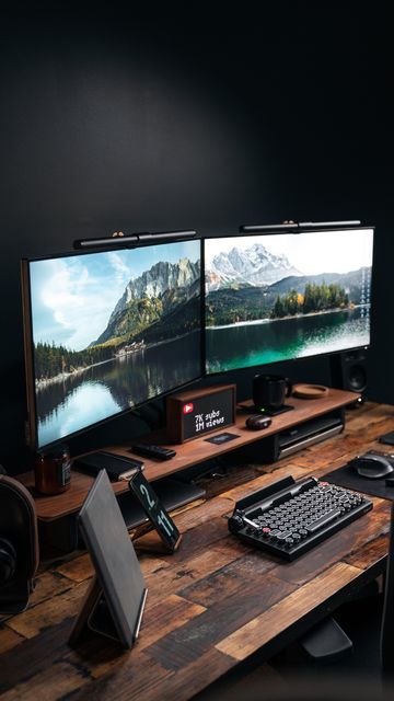 two computer monitors sitting next to each other on a wooden desk in front of a black wall