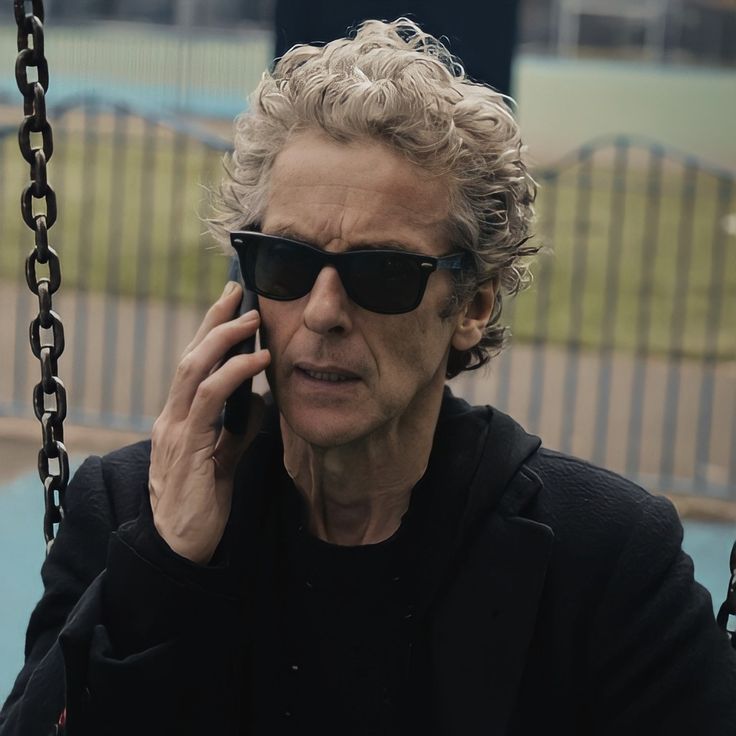 an older man wearing sunglasses talking on a cell phone while sitting on a swingset