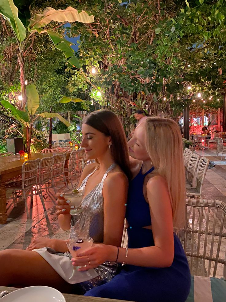 two beautiful young women sitting next to each other at a table with wine glasses in their hands