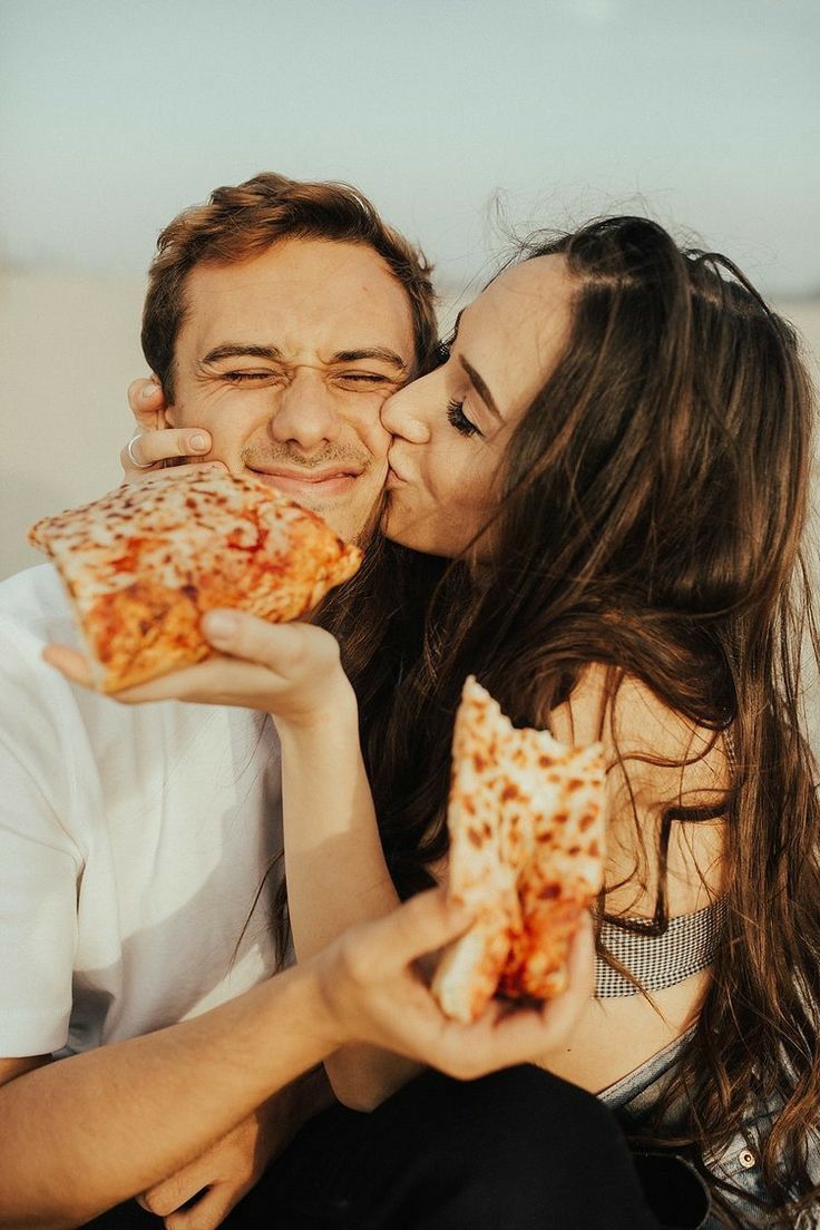 a man and woman sharing a slice of pizza on top of each other in front of the ocean