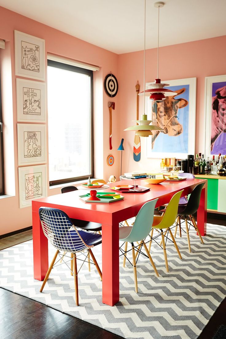 a dining room table with colorful chairs and artwork on the wall in the back ground