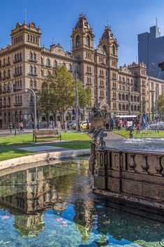 the fountain in front of the building is surrounded by tall buildings and green grass on both sides