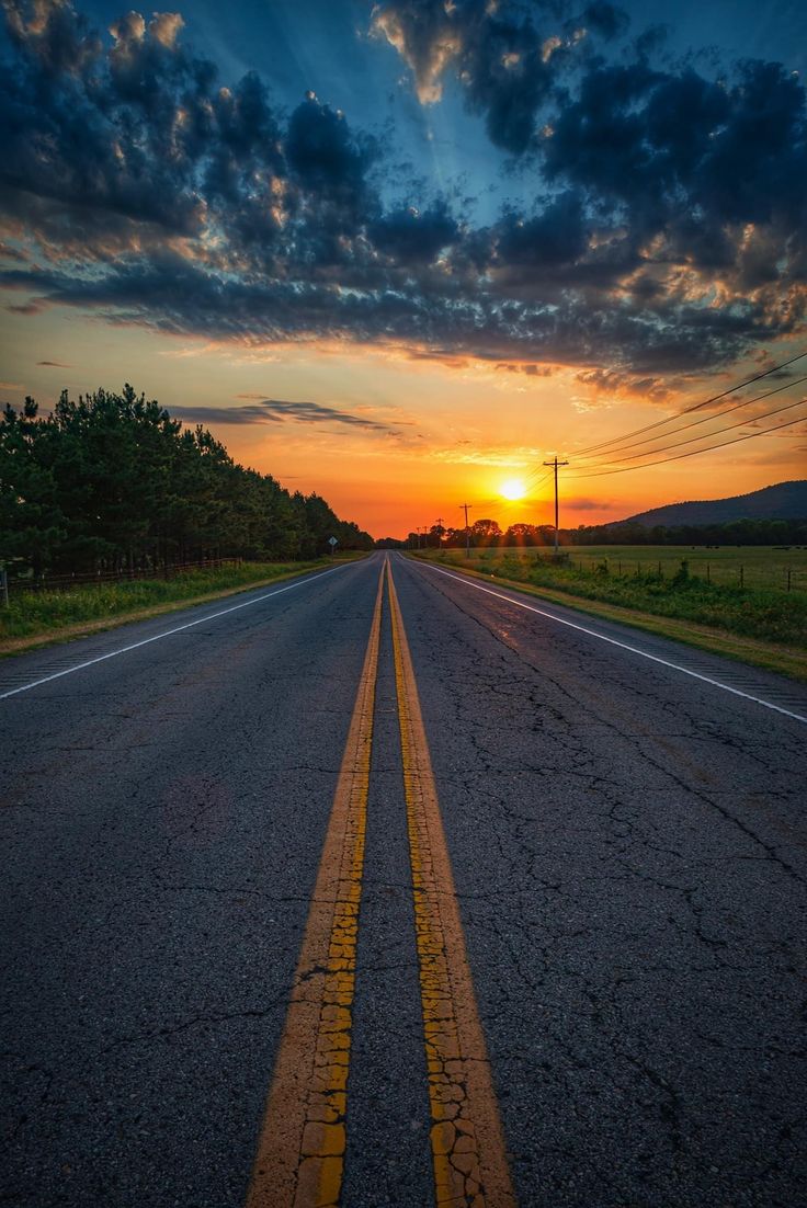 the sun is setting on an empty road with yellow lines in the middle and trees to the side