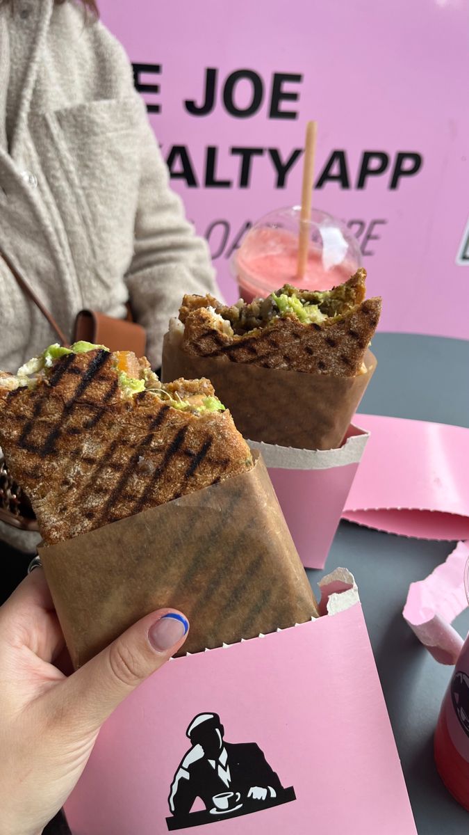 two sandwiches in paper wrappers sitting on a table next to pink cups with drinks