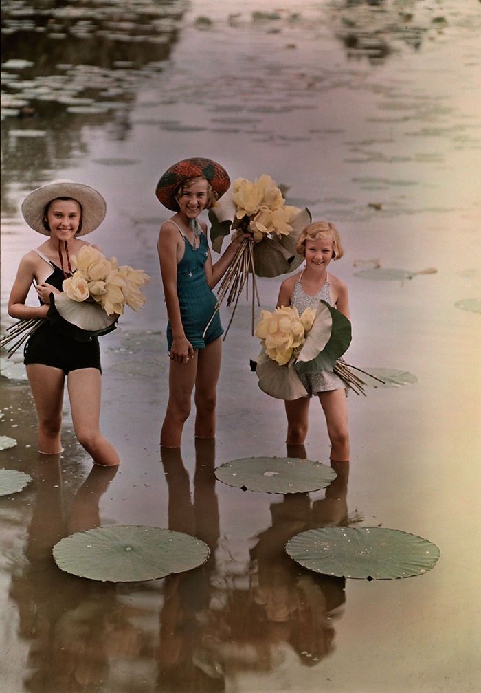 three women in swimsuits and hats standing on the beach with water lillies