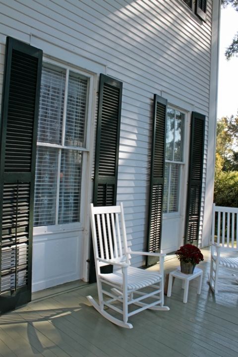 two white rocking chairs sitting on the front porch of a house with shutters open