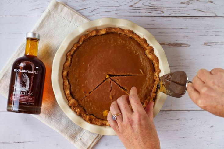 a person cutting into a pie on top of a white wooden table next to a bottle of booze