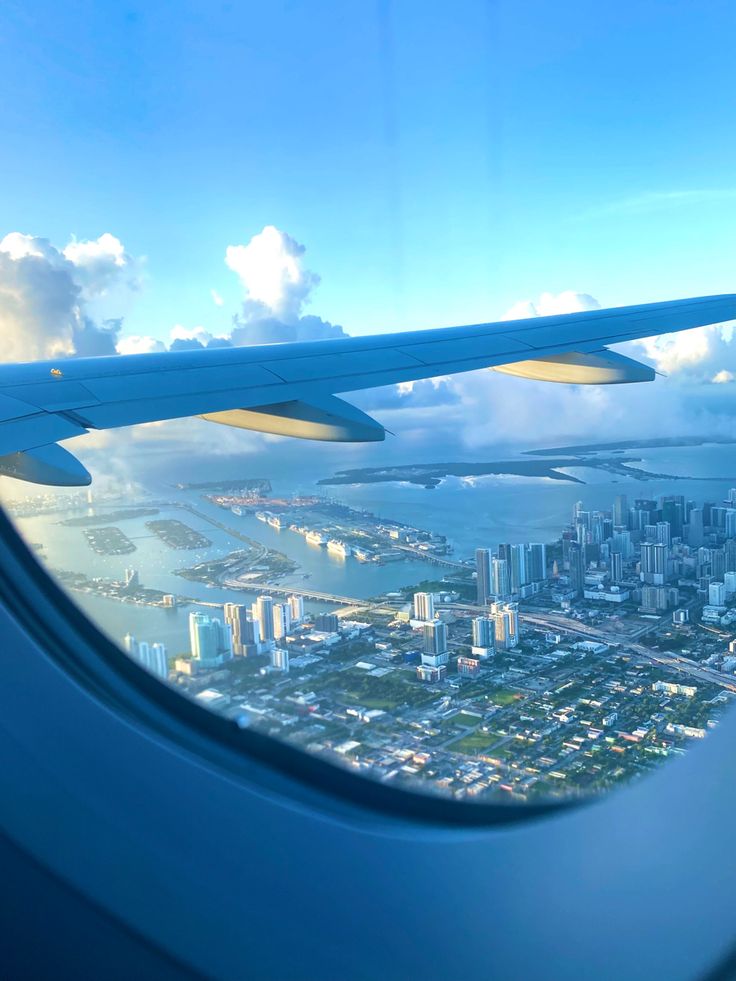 the view from an airplane window looking down on a city and water below it, with clouds in the sky