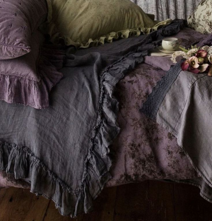 a bed covered in purple ruffled sheets and pillows next to a vase with flowers