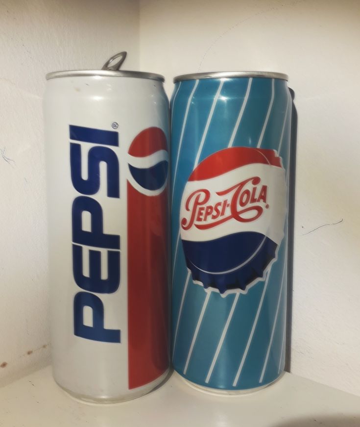 two pepsi cola cans sitting next to each other on a shelf in a room with white walls