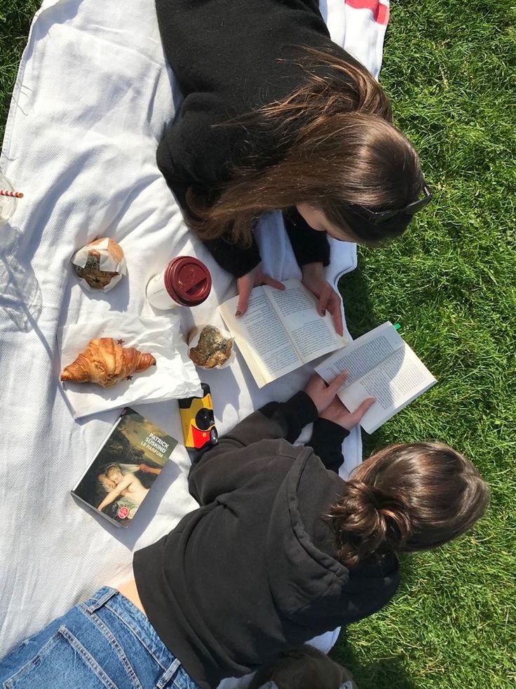 two people sitting on the grass reading books and eating croissants while looking at pictures