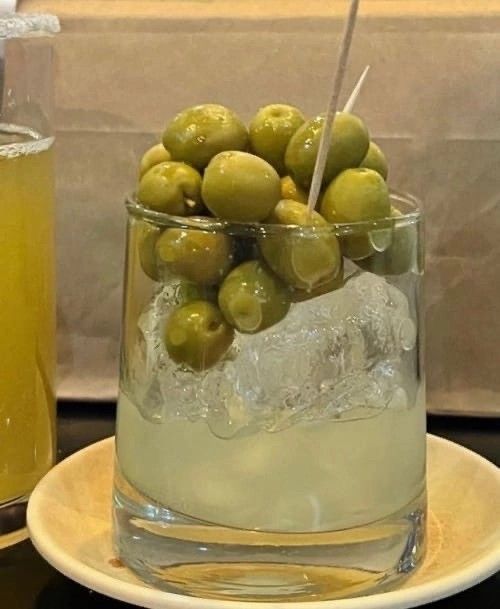 a glass filled with green olives on top of a plate next to a drink