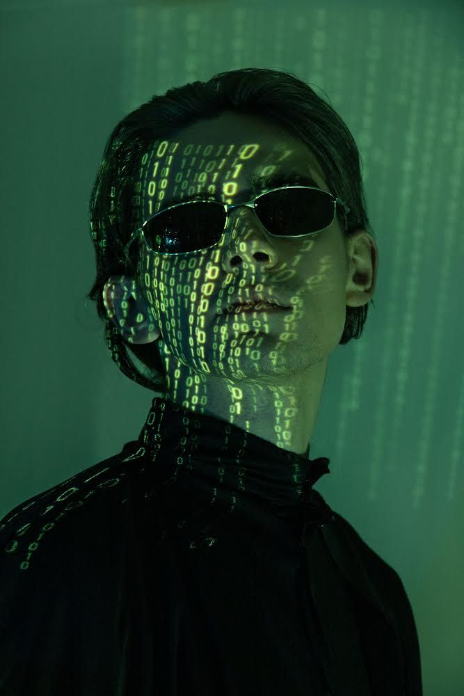 a man wearing sunglasses with numbers projected on his face in the back ground and behind him is a green background