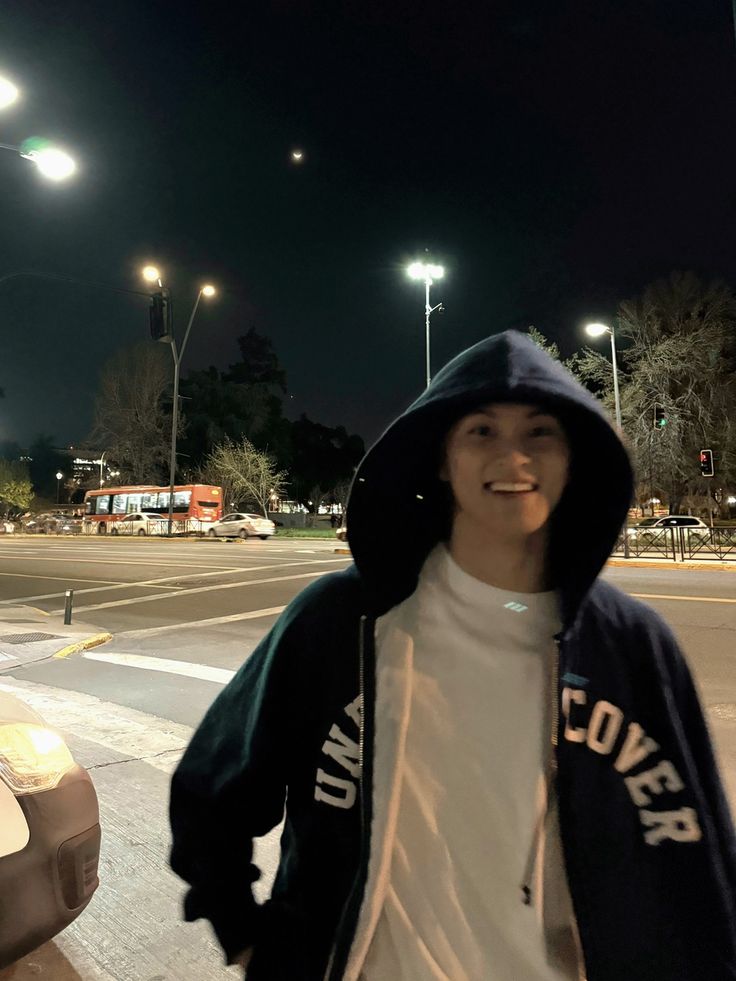 a man in a hoodie is standing on the sidewalk at night with his hands behind his back