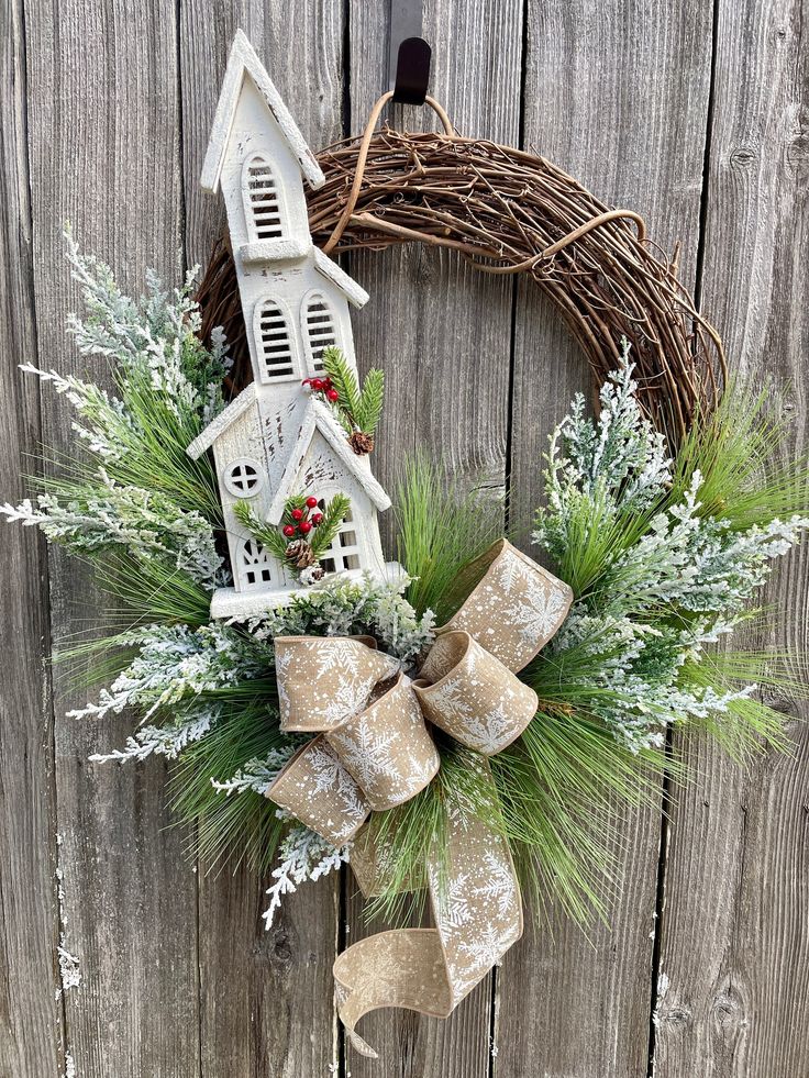 a wreath with a church and evergreens hanging on the side of a wooden fence