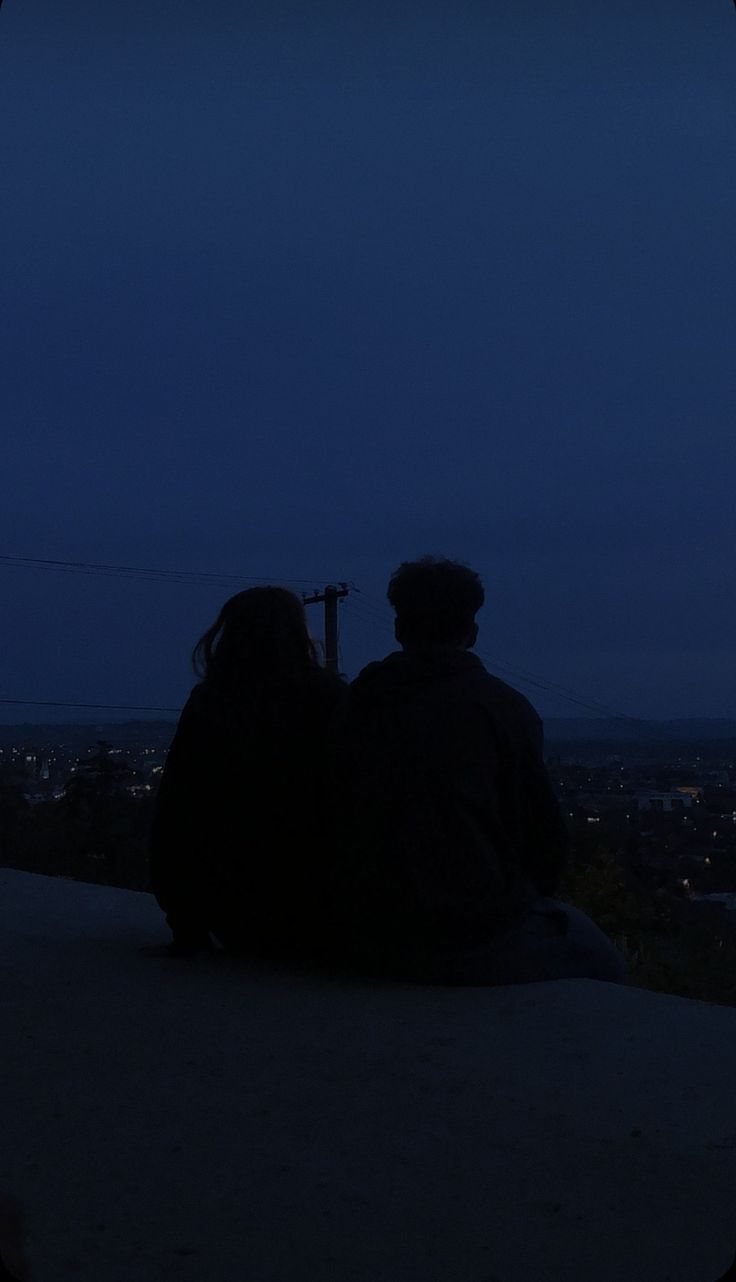 two people are sitting on a ledge at night looking out into the city below them