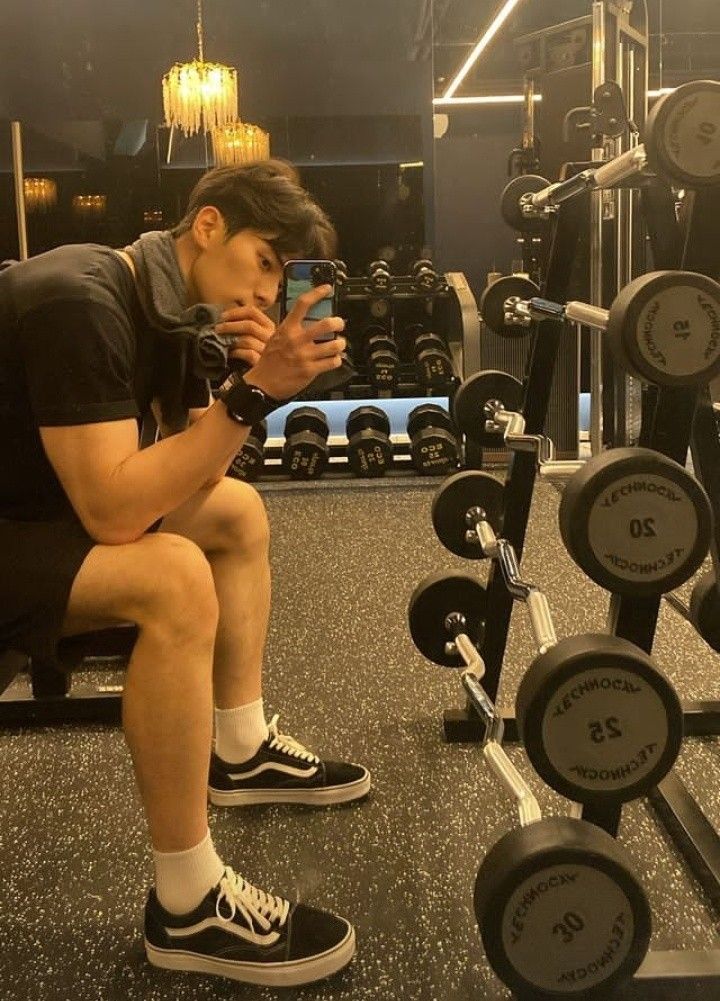 a man squatting down while looking at his cell phone in a gym with rows of dumbbells