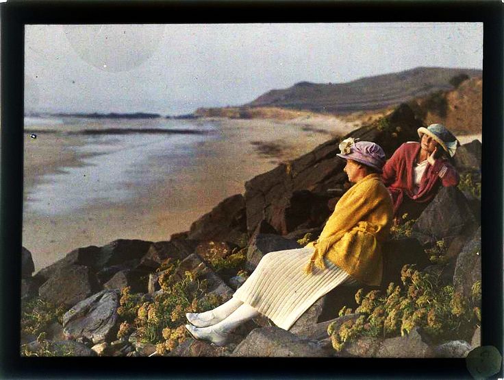 two women sitting on rocks looking out at the ocean and mountains in an old fashion photo