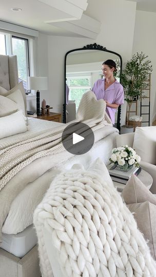 a woman standing next to a bed in a room with white furniture and pillows on it