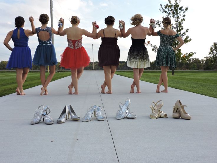 four women in dresses and high heels standing on the sidewalk with their arms around each other