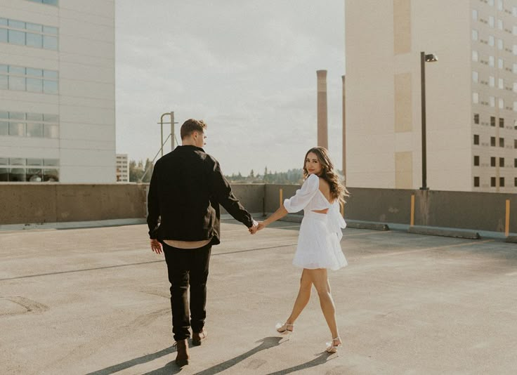 a man and woman holding hands walking across an empty parking lot in front of tall buildings