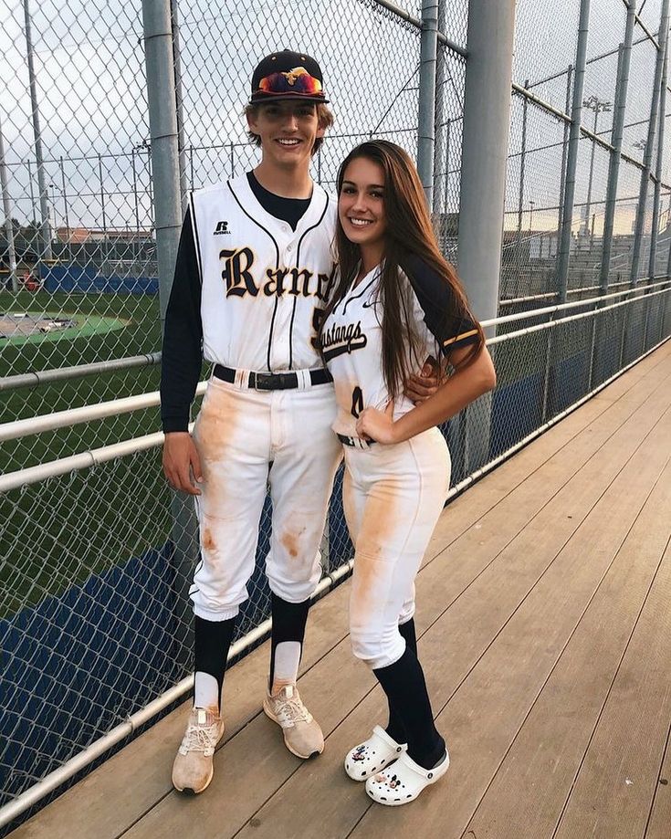 a man and woman in baseball uniforms posing for a photo on a wooden deck next to a fence