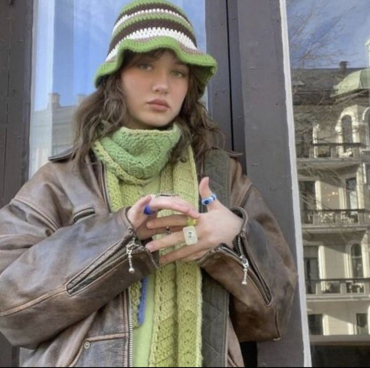 a woman wearing a green hat and scarf standing in front of a window with her hand on her hip