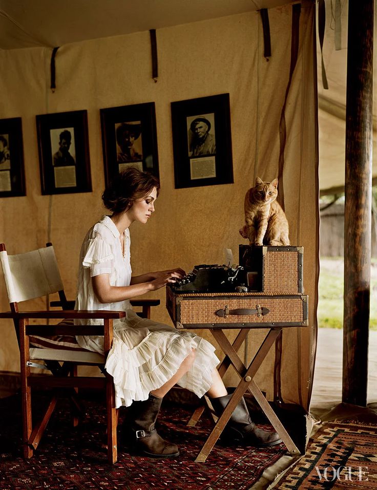 a woman sitting at a table typing on an old typewriter with a cat standing next to her