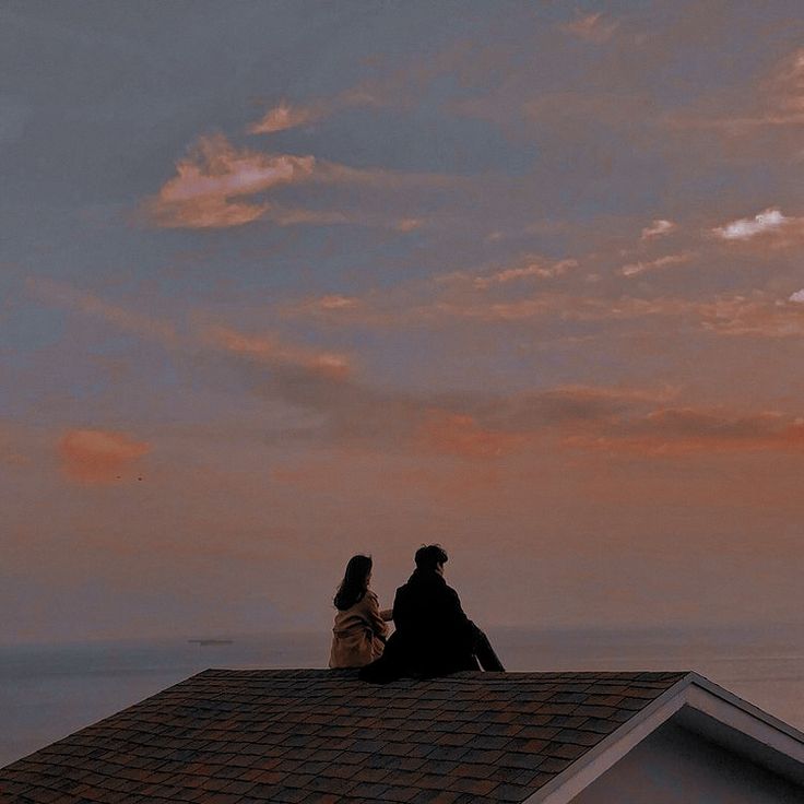 two people sitting on top of a roof looking out at the ocean under a cloudy sky