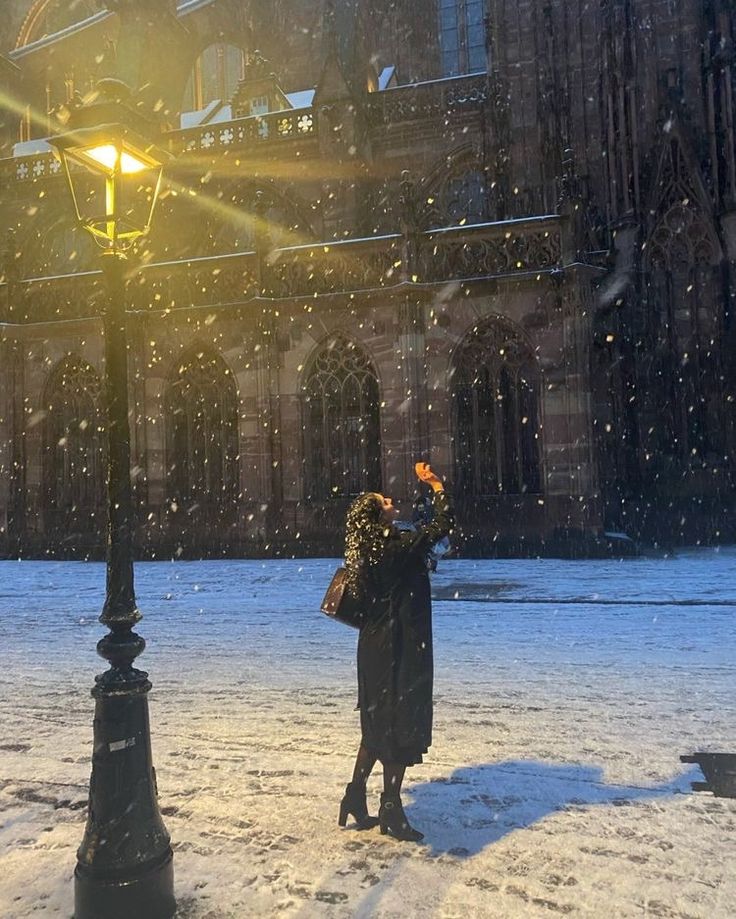 a woman standing in the snow next to a street light and lamp post with her hand up