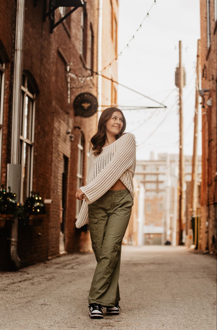 a woman is standing in an alley way wearing green pants and a white knitted sweater