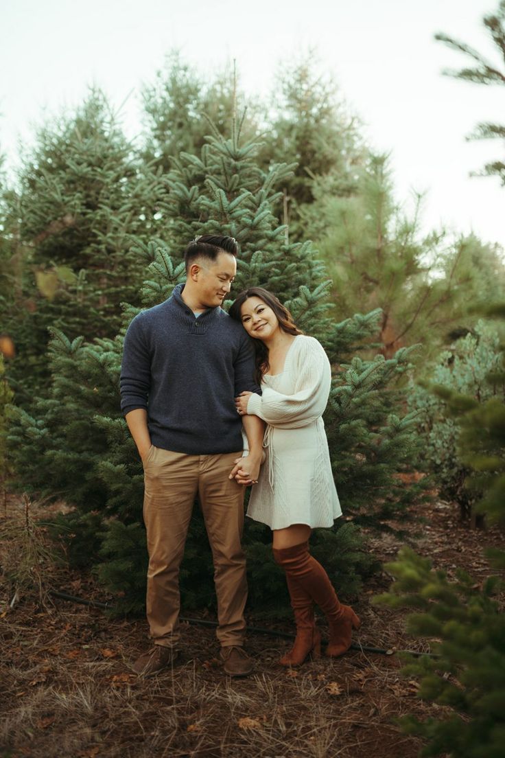 a man and woman standing in front of a christmas tree at an evergreen forest with their arms around each other