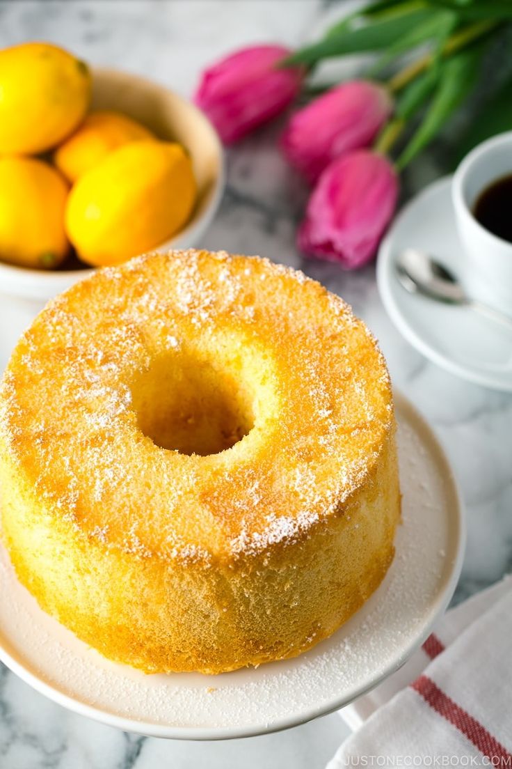 a lemon cake on a plate next to some flowers and two mugs with coffee