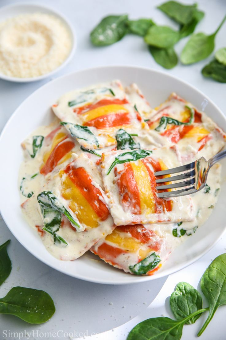 a white plate topped with ravioli covered in cheese and sauce next to spinach leaves