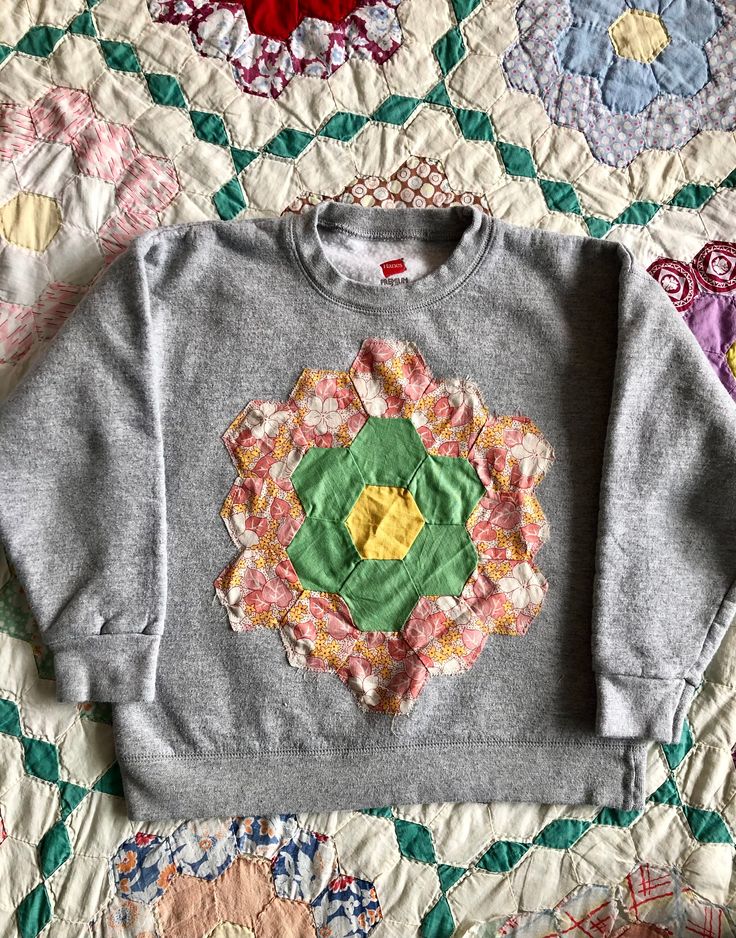 a gray sweater with an embroidered flower on the front and bottom, sitting on a quilted surface