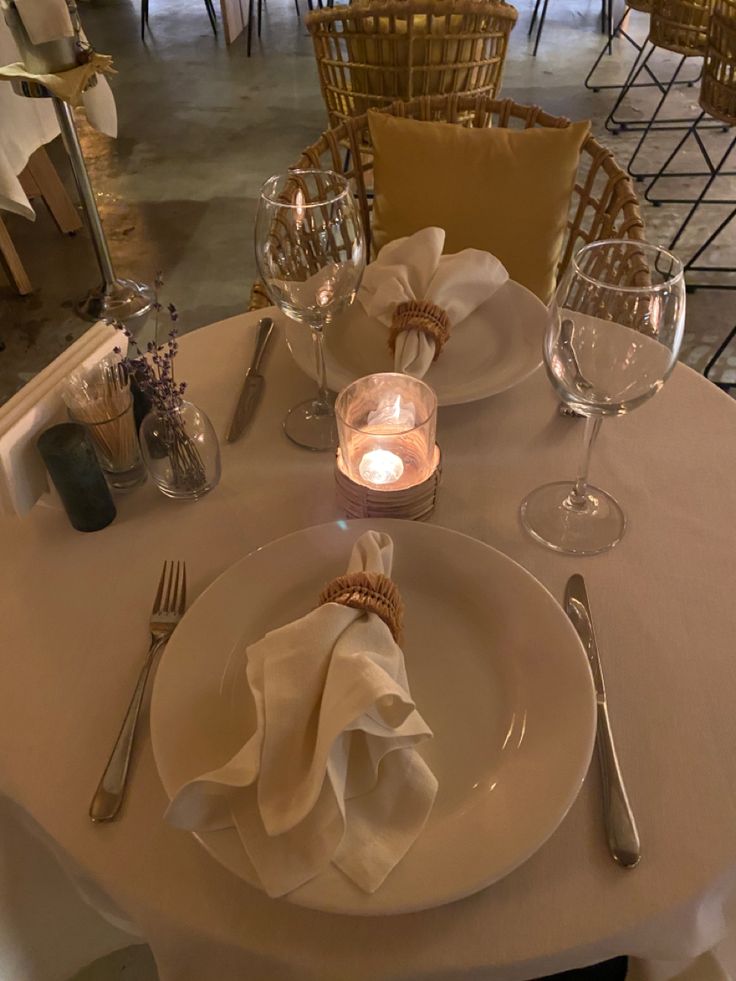 the table is set for dinner with white linens and silverware, candles and napkins