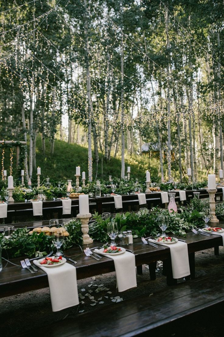 an outdoor wedding setup with long tables and white linens on the table, surrounded by tall trees