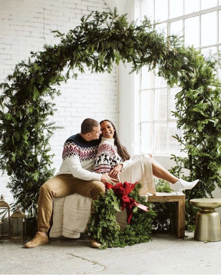 a man and woman sitting on a bench in front of a christmas wreath with greenery