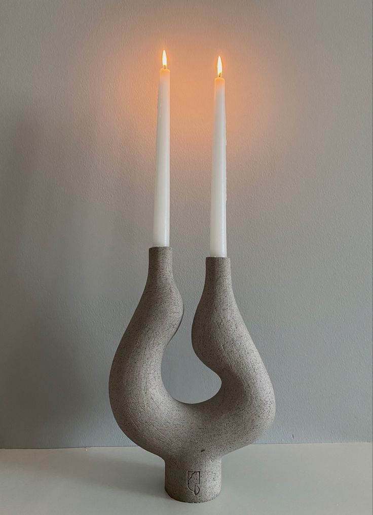 two white candles are sitting in an unusual shaped vase on a table with grey walls behind them