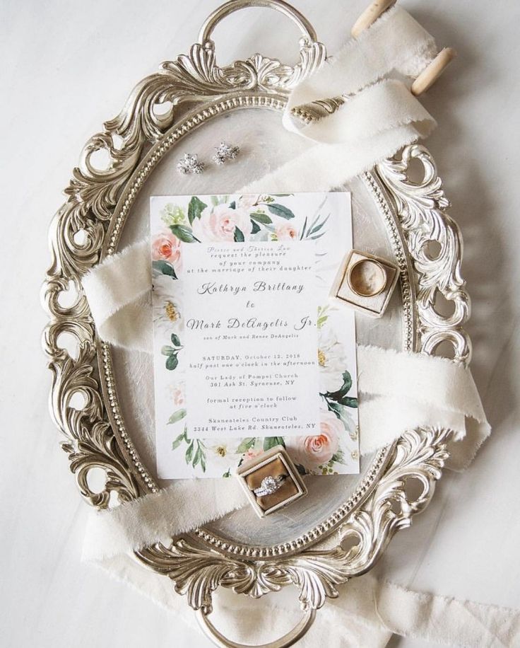 a wedding card and ring on a silver frame with white ribbon around it, surrounded by flowers