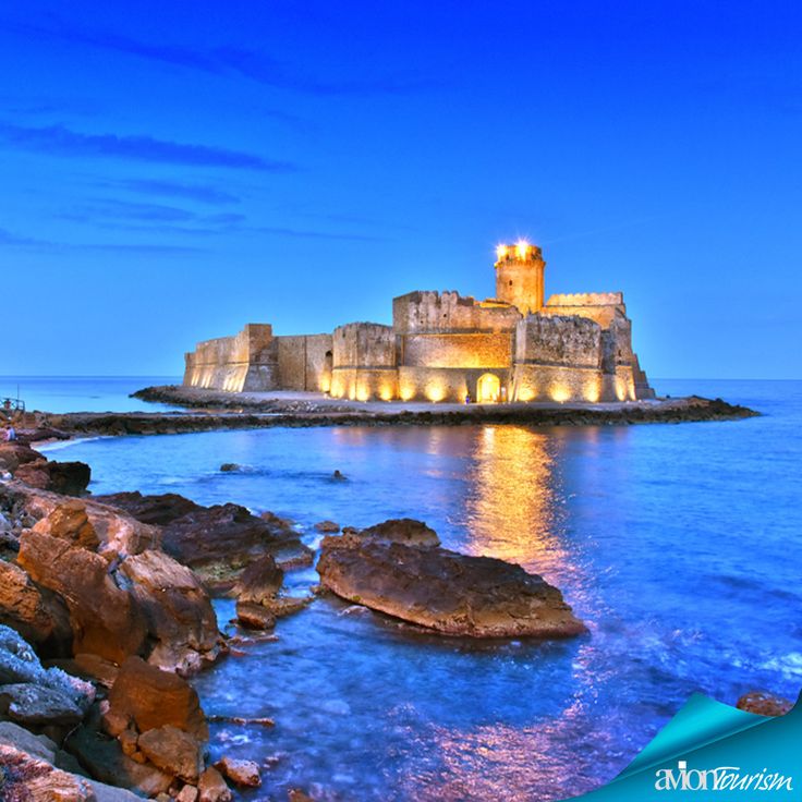 the castle is lit up at night by the water's edge with rocks in front of it