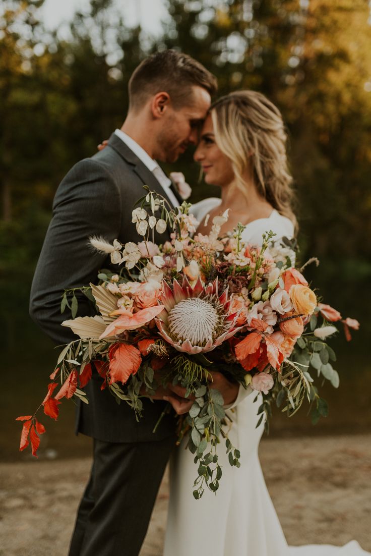 a bride and groom standing together in front of some trees with orange flowers on their bouquets