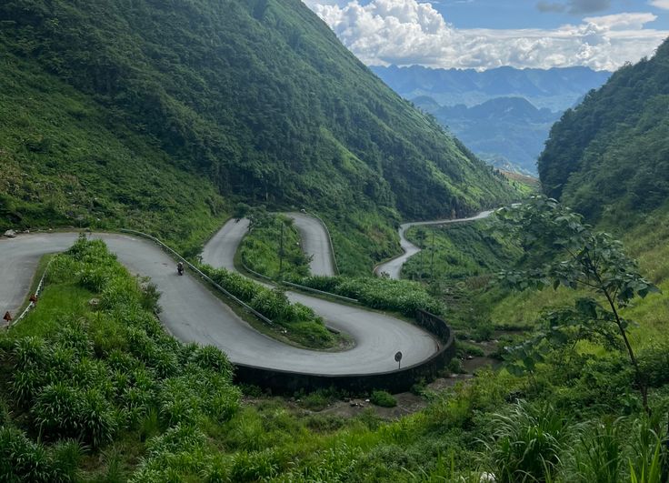 the winding road is surrounded by mountains and greenery on both sides, as well as two bikers riding down it