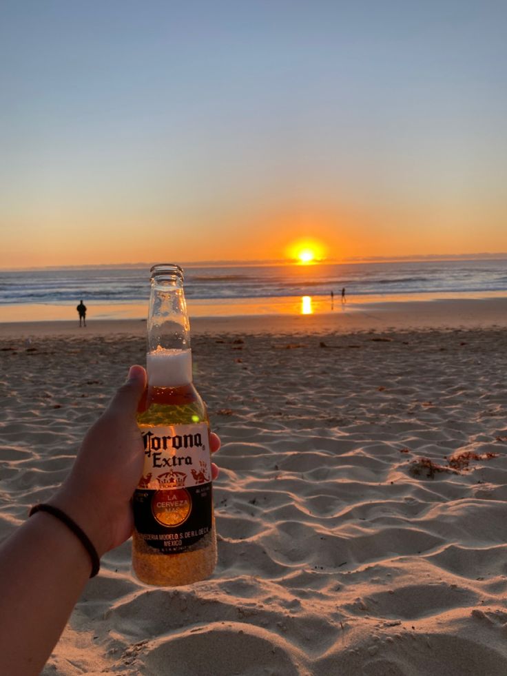 a person holding up a beer bottle on the beach at sunset with people in the background